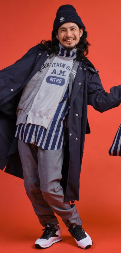 A man wearing a Mountain Research outfit featuring a double zip hoodie, navy blue overcoat and grey pants