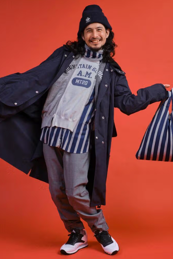 A man wearing a Mountain Research outfit featuring a double zip hoodie, navy blue overcoat and grey pants