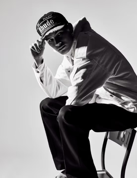A man sitting on a chair wearing a Rhude Racing hat, button up jacket and black pants