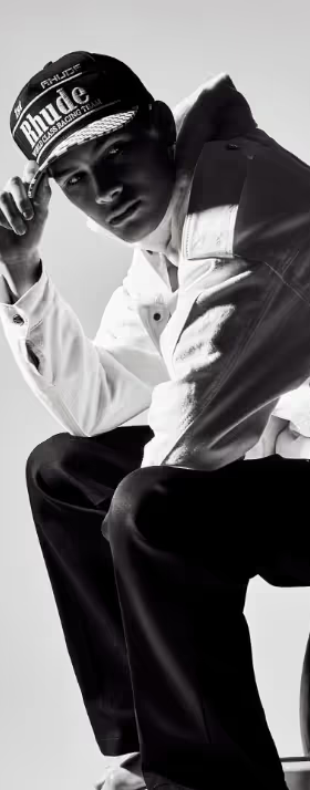 A man sitting on a chair wearing a Rhude Racing hat, button up jacket and black pants