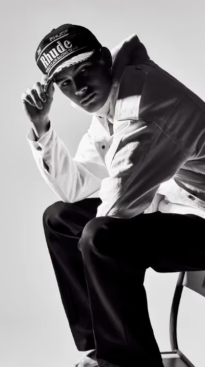 A man sitting on a chair wearing a Rhude Racing hat, button up jacket and black pants
