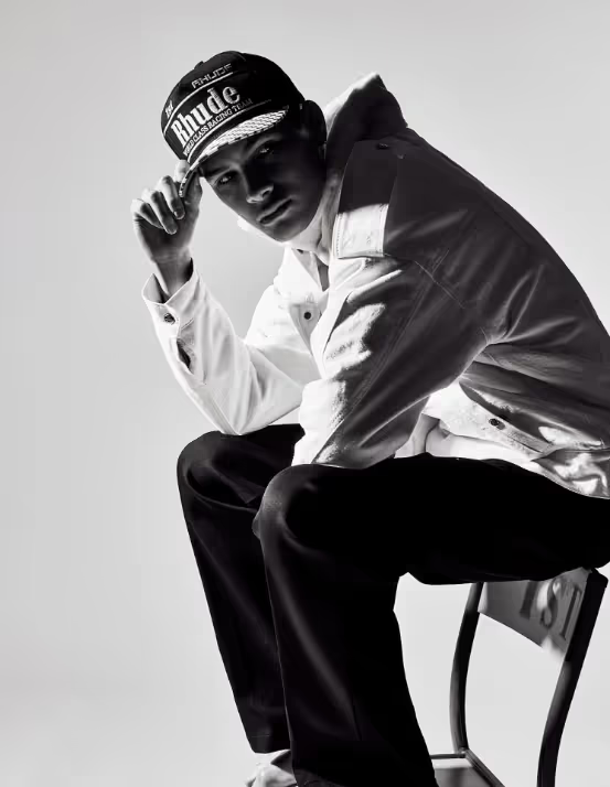 A man sitting on a chair wearing a Rhude Racing hat, button up jacket and black pants