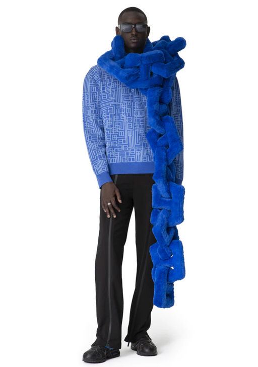 A man wearing a Piéces Uniqes outfit feauring an oversize blue pillow-like scarf and navy blue pants