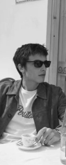 A man wearing a Harmony Paris t-shirt, jacket and sunglasses while having a cappucino