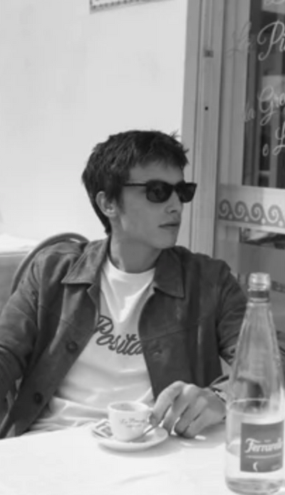 A man wearing a Harmony Paris t-shirt, jacket and sunglasses while having a cappucino