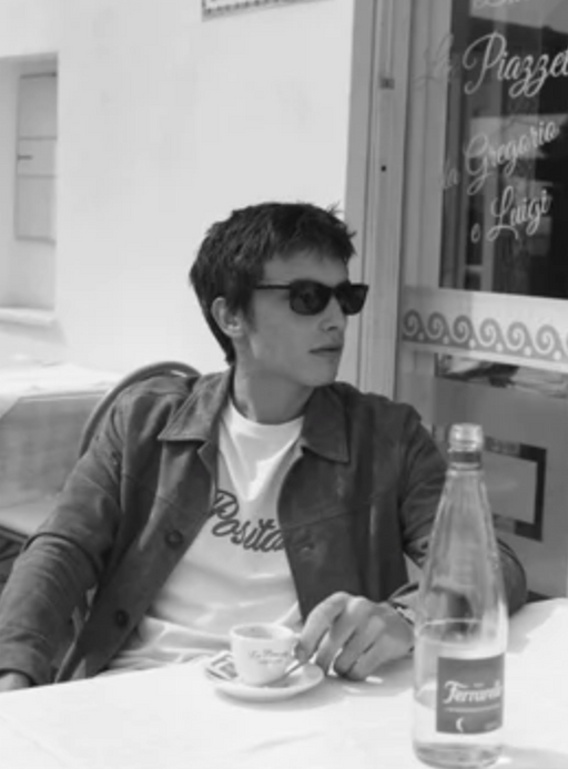 A man wearing a Harmony Paris t-shirt, jacket and sunglasses while having a cappucino
