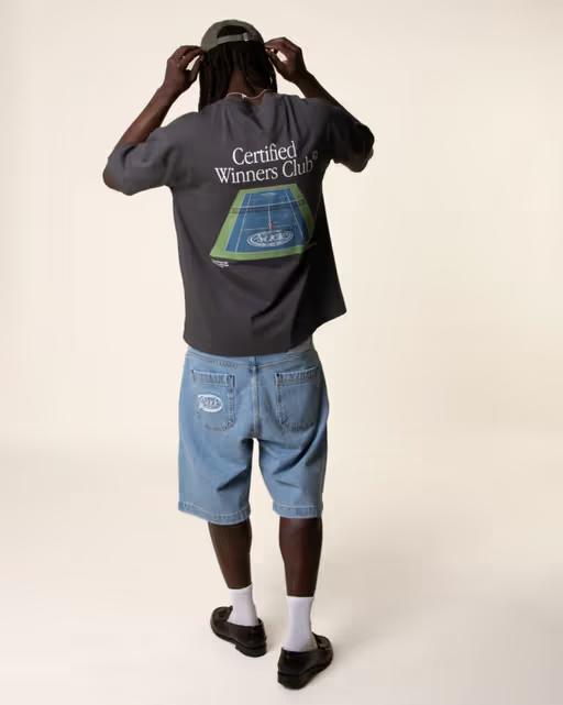 A man wearing a Nude Project tennis t-shirt with denim shorts