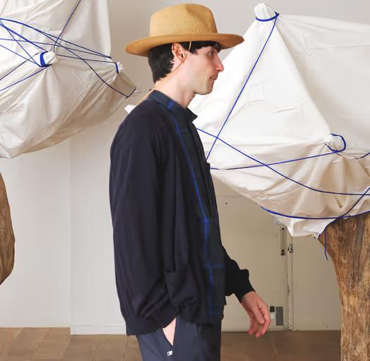 A man wearing a Nonnative navy blue sweater, blue button up and brown flat brimmed hat
