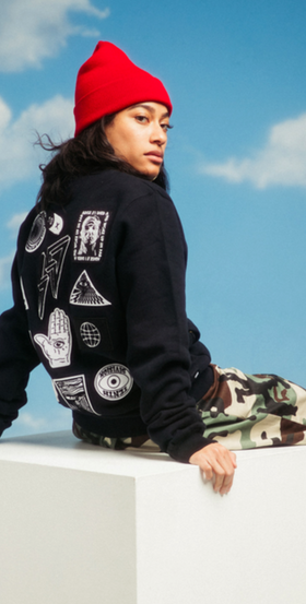 A woman sitting and wearing a 10 Deep hoodie with camo pants and a red beanie