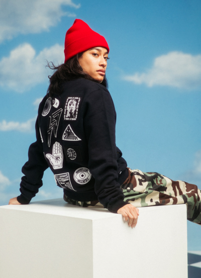 A woman sitting and wearing a 10 Deep hoodie with camo pants and a red beanie