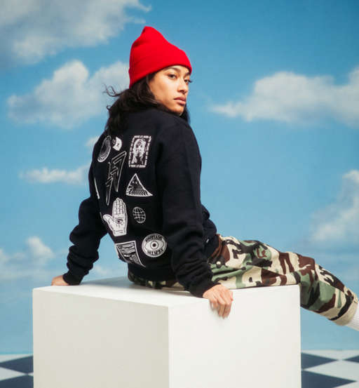 A woman sitting and wearing a 10 Deep hoodie with camo pants and a red beanie