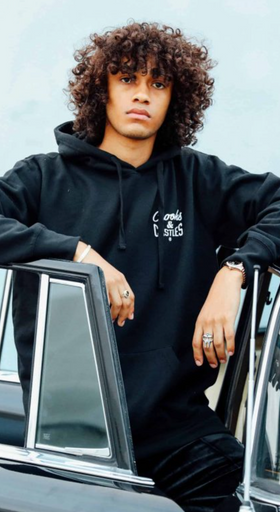 A man wearing a Crooks & Castles hoodie standing beside a black car