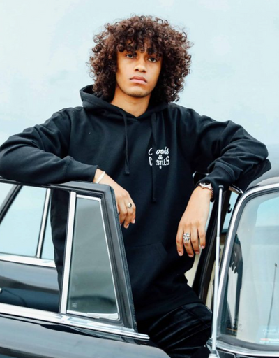 A man wearing a Crooks & Castles hoodie standing beside a black car