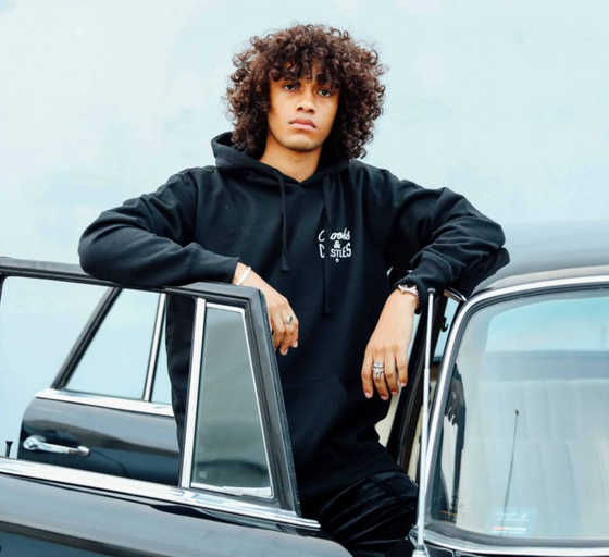 A man wearing a Crooks & Castles hoodie standing beside a black car