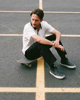 A man sitting on his skateboard wearing a white Globe t-shirt and black pants