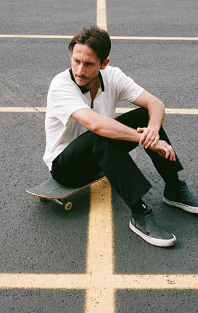 A man sitting on his skateboard wearing a white Globe t-shirt and black pants