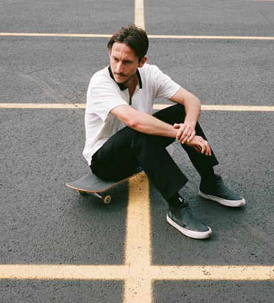 A man sitting on his skateboard wearing a white Globe t-shirt and black pants