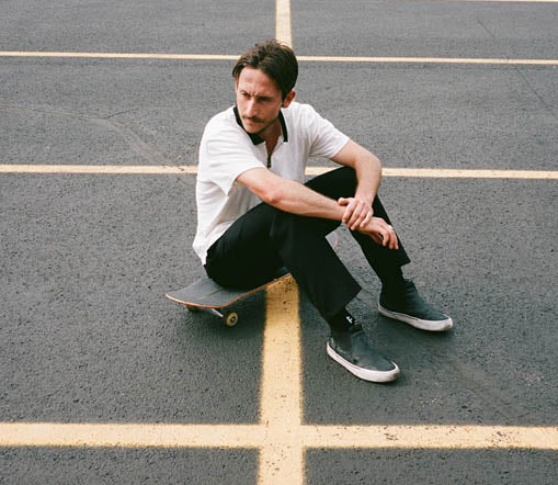 A man sitting on his skateboard wearing a white Globe t-shirt and black pants