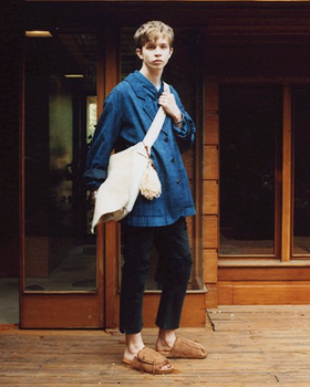 A man wearing a Visvim denim jacket, beige carryall, dark pants and brown slippers