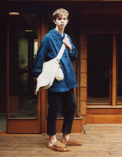 A man wearing a Visvim denim jacket, beige carryall, dark pants and brown slippers