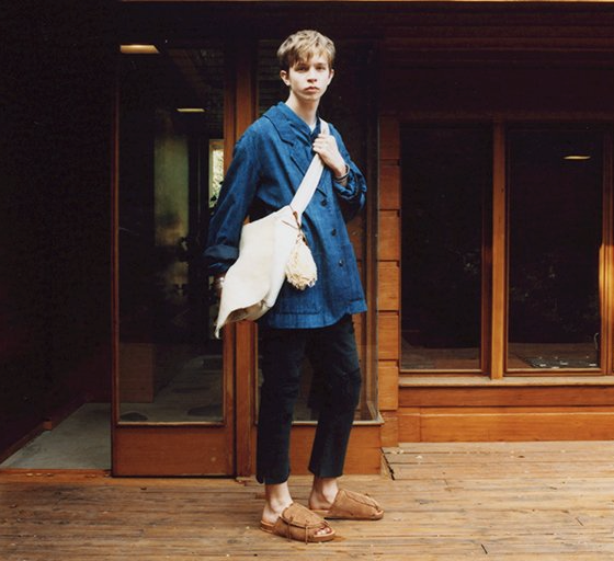 A man wearing a Visvim denim jacket, beige carryall, dark pants and brown slippers