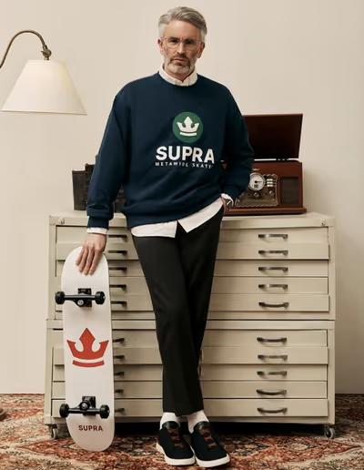 A man wearing a Supra crewneck sweater with a white button shirt underneath