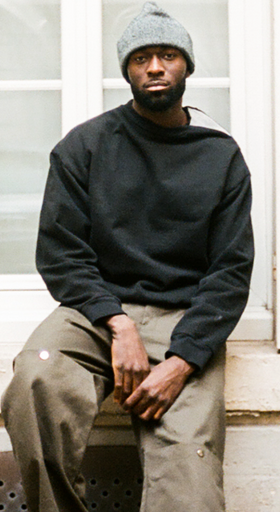 A man wearing a Spencer Badu black sweater and dark green pants