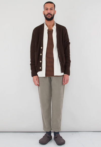 A man wearing a Folk Clothing layered outfit with a brown cardigan and white button up and grey pants
