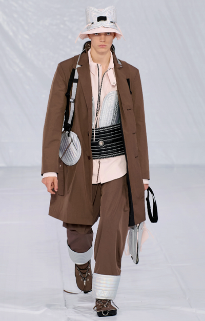 A man walking a runway in a Craig Green designer construction-influenced brown outfit with an overcoat and wide pants