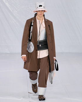 A man walking a runway in a Craig Green designer construction-influenced brown outfit with an overcoat and wide pants