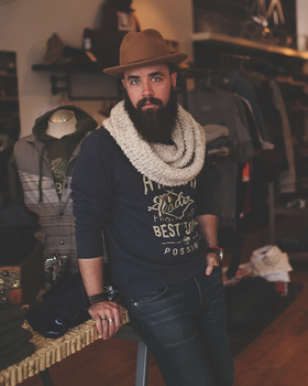 A man wearing a James Dant outfit featuring a brown hat, cream scarf and navy blue branded long-sleeve t-shirt