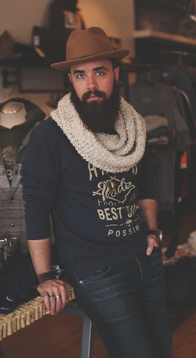 A man wearing a James Dant outfit featuring a brown hat, cream scarf and navy blue branded long-sleeve t-shirt