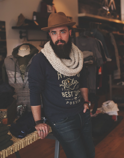 A man wearing a James Dant outfit featuring a brown hat, cream scarf and navy blue branded long-sleeve t-shirt