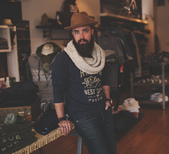 A man wearing a James Dant outfit featuring a brown hat, cream scarf and navy blue branded long-sleeve t-shirt