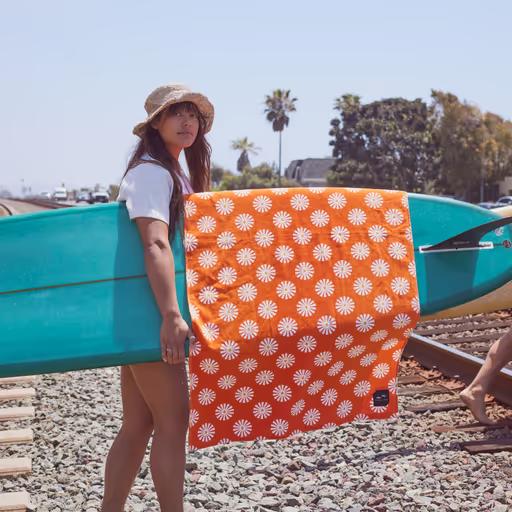 A woman holding a surfboard with an orange slowtide towel draped over it