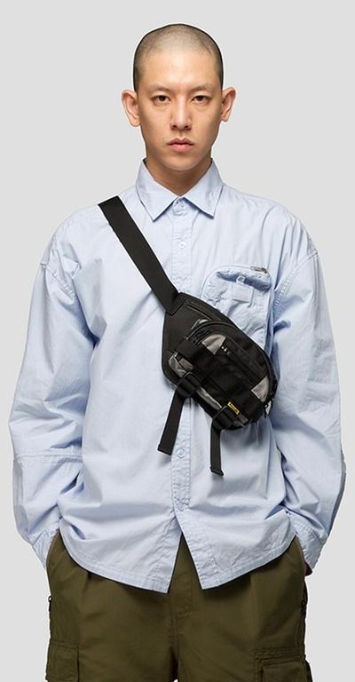 A man wearing a 4DIMENSION outfit featuring a light blue button up and a black bag slinged across chest