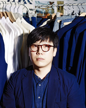 A man wearing a Document blue button up and cardigan surrounded by button up shirts