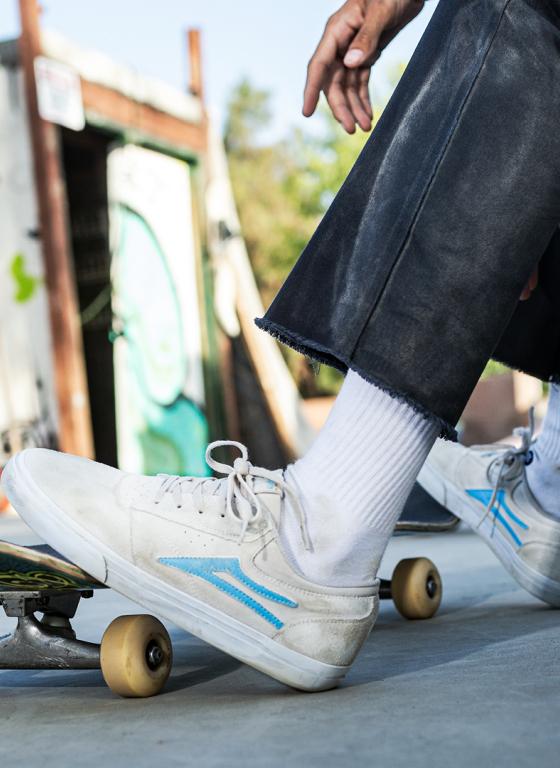 A man skateboarding wearing a cream pair of Lakai shoes with a baby blue logo
