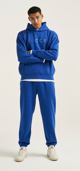A man wearing a light beige Nicce Logo hoodie with denim shorts