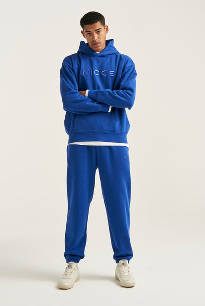 A man wearing a light beige Nicce Logo hoodie with denim shorts