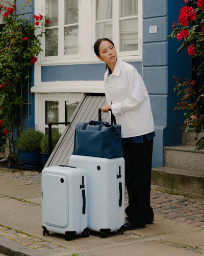 A lady holding light blue Monos roller luggage outside the front door