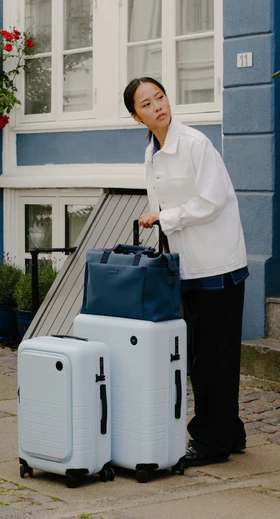 A lady holding light blue Monos roller luggage outside the front door
