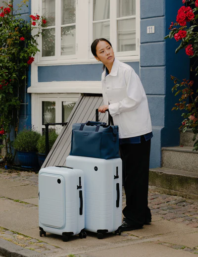 A lady holding light blue Monos roller luggage outside the front door