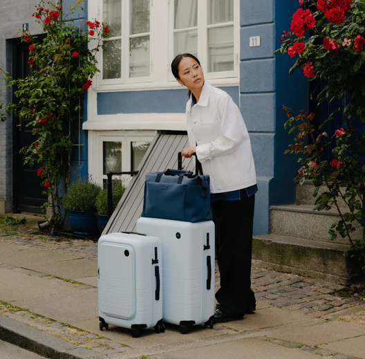 A lady holding light blue Monos roller luggage outside the front door