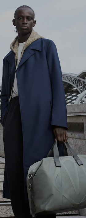 A man holding a Rimowa grey bag and a blue trench coat