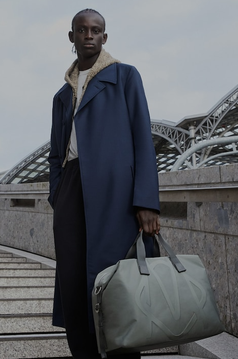A man holding a Rimowa grey bag and a blue trench coat