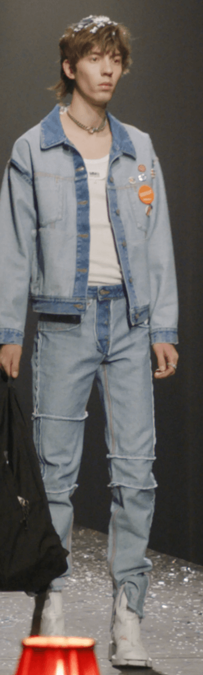 A man on a fashion runway wearing a Maison Margiela denim jacket and matching pants along with a white high-heel boots