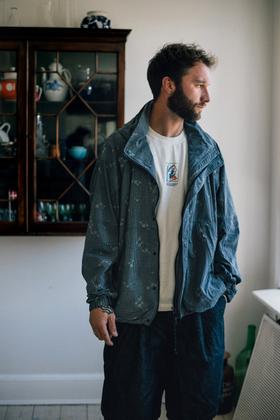 A man wearing a blue-grey printed 18 East windbreaker over a white embroidered tee and dark trousers