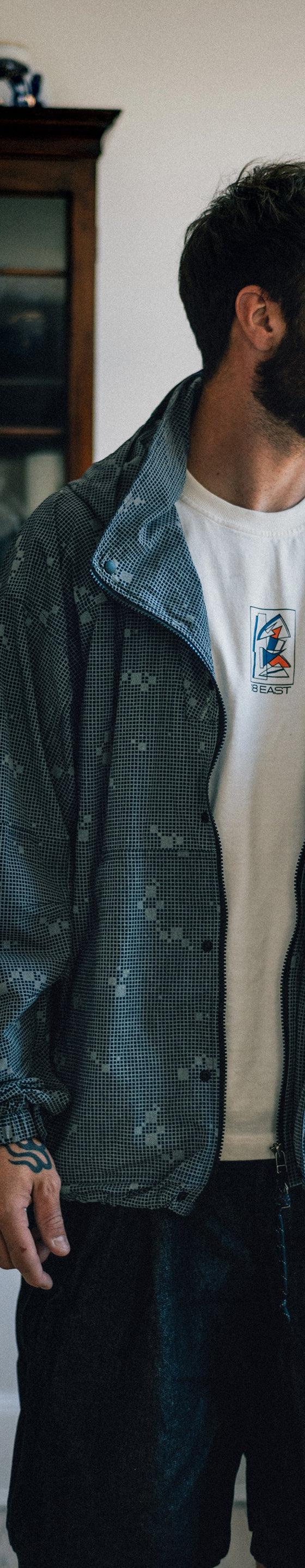 A man wearing a blue-grey printed 18 East windbreaker over a white embroidered tee and dark trousers