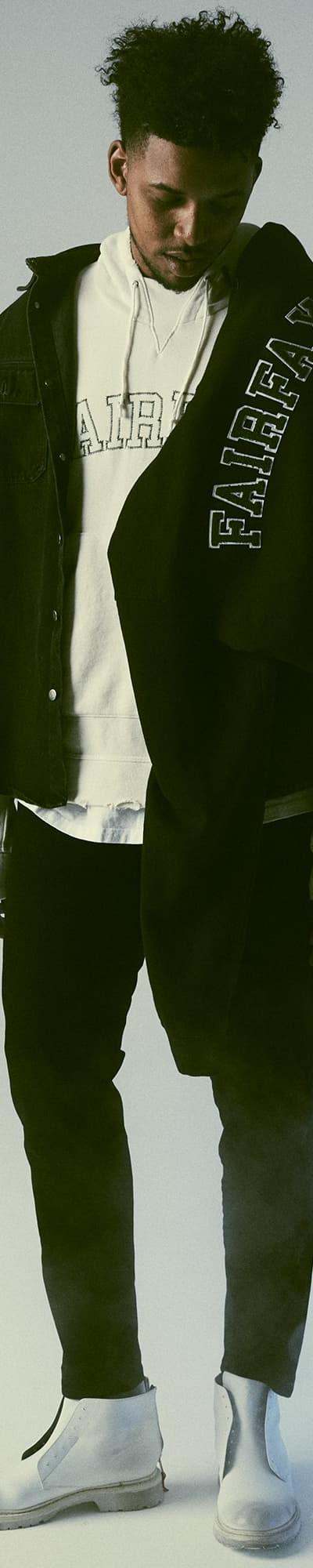 A man wearing a black 424 Fairfax overshirt layered over a white FAIRFAX hoodie with black trousers and white boots